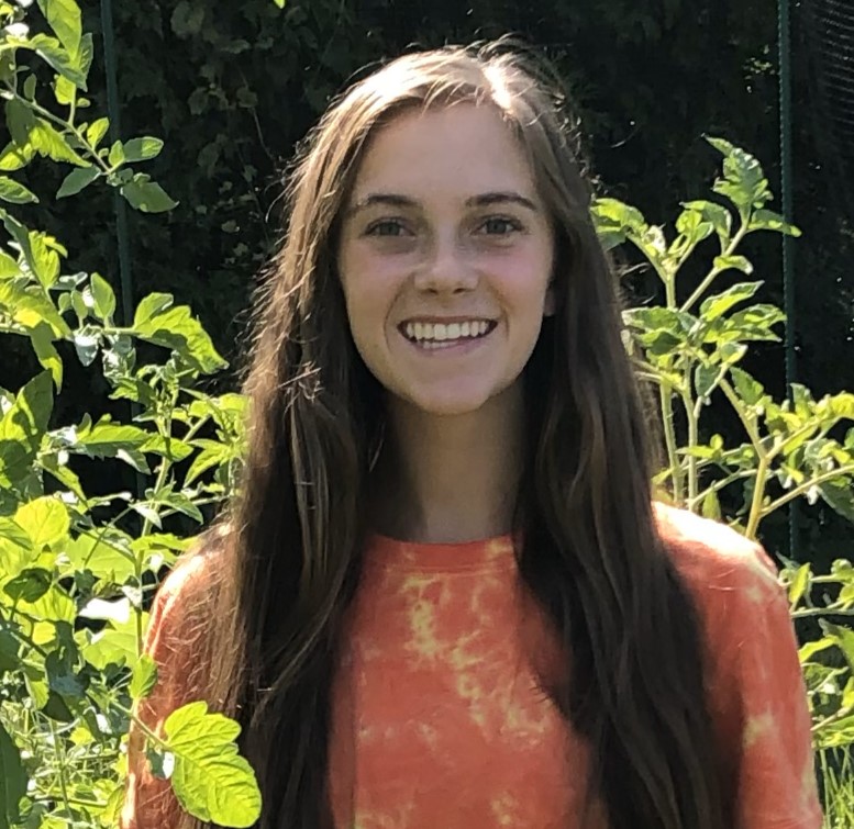 Sarah DeVito, a white, brown haired, blue eyed woman wearing an orange and yellow tie-dye shirt standing among plants.